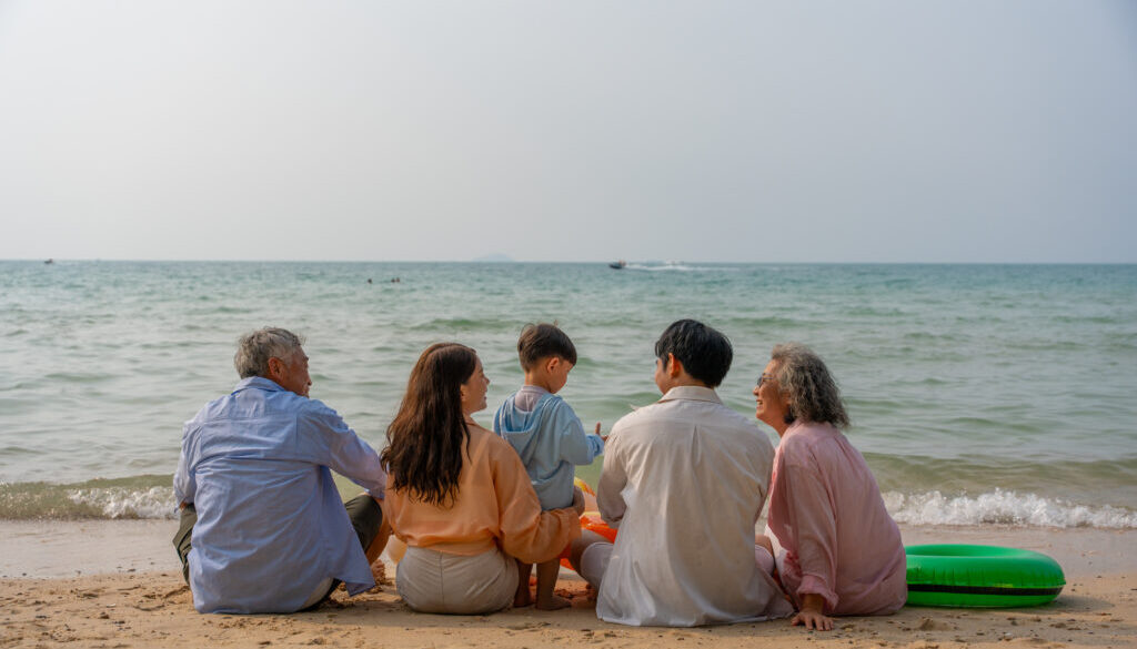 Happy multi-generational Asian family sitting on the beach with their backs to the camera, raising hands and enjoying fun vacation moment together by the sea. family, beach, vacation, summer. elderly