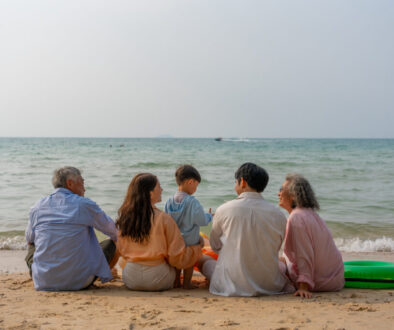 Happy multi-generational Asian family sitting on the beach with their backs to the camera, raising hands and enjoying fun vacation moment together by the sea. family, beach, vacation, summer. elderly