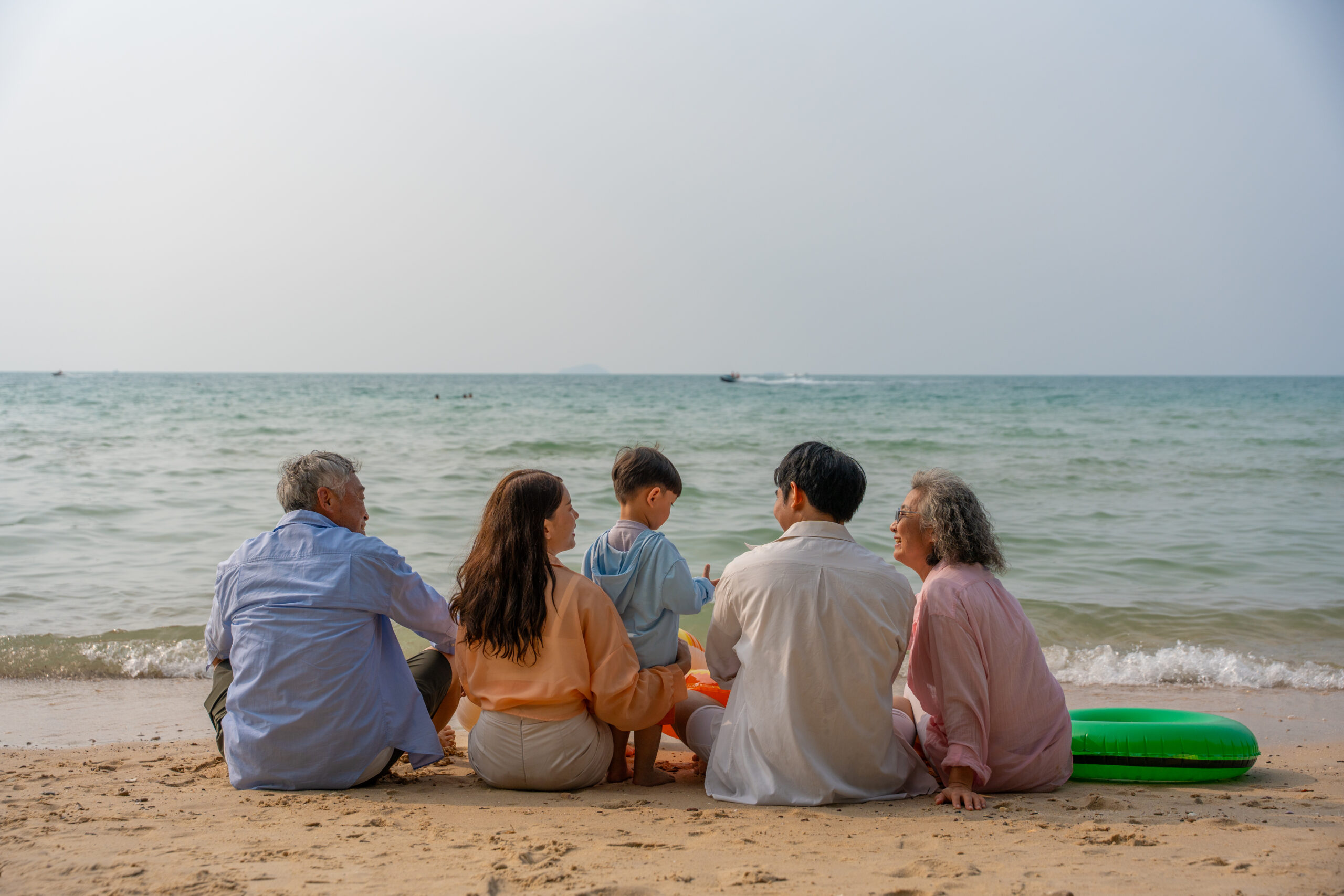 Happy multi-generational Asian family sitting on the beach with their backs to the camera, raising hands and enjoying fun vacation moment together by the sea. family, beach, vacation, summer. elderly