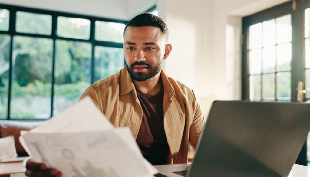 Man, documents and home with laptop for financial statistics, report or performance review. Male person, freelancer or remote worker with paperwork on computer for finance, expenses or bills at house