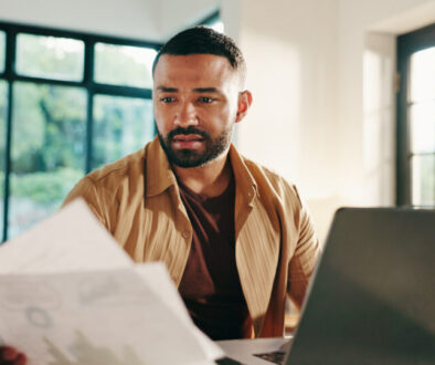 Man, documents and home with laptop for financial statistics, report or performance review. Male person, freelancer or remote worker with paperwork on computer for finance, expenses or bills at house
